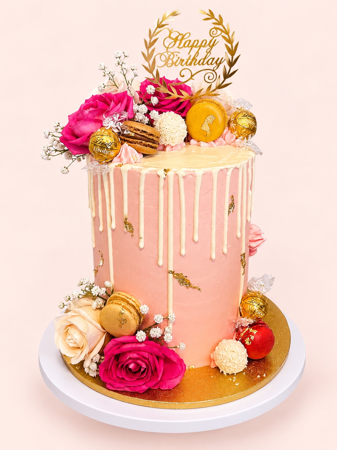 Tall pink drip cake with pink and cream roses, gold macarons, chocolates, gold leaf and a gold Happy Birthday topper