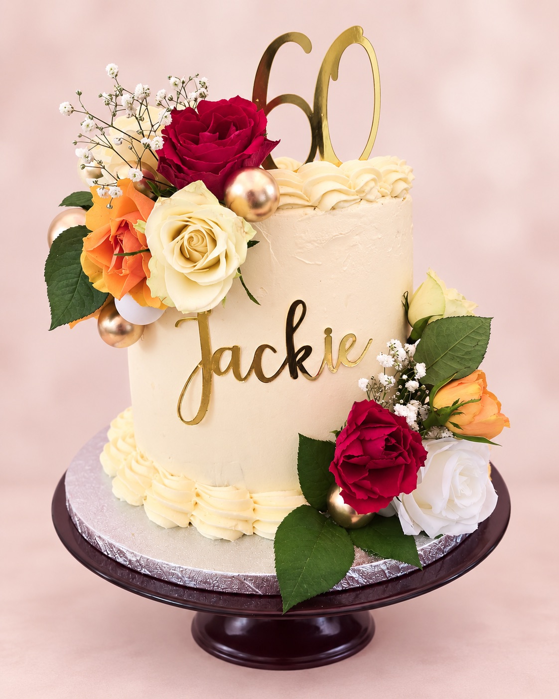 Tall cream buttercream cake with “Jackie” and a gold 60 topper, decorated with red, orange and cream roses and gold balls