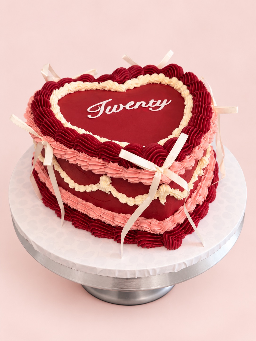 Red heart-shaped vintage cake with pink and cream piping, cream ribbon bows and “Twenty” piped on top